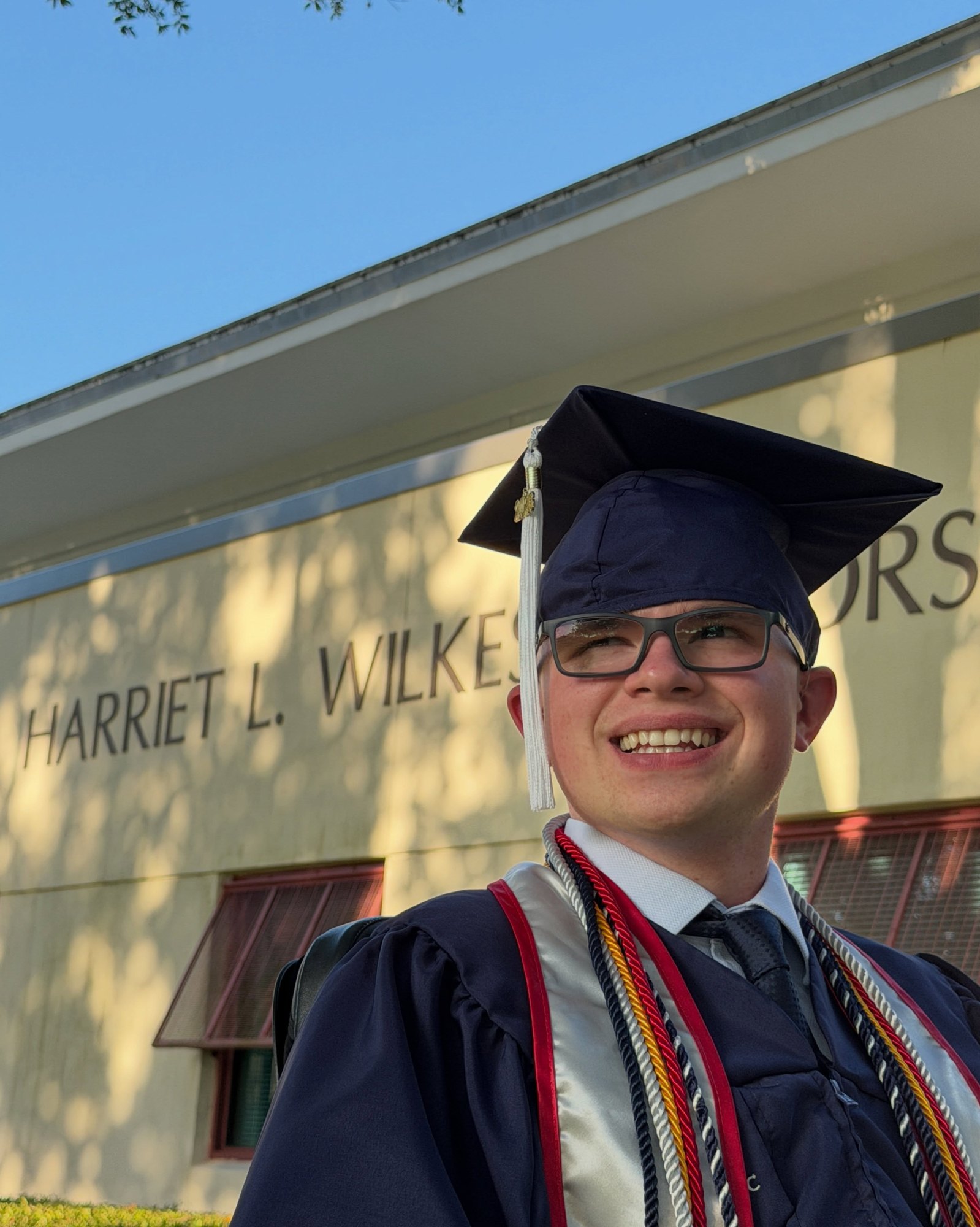 Ryleigh Newman in his graduation cap and gown in his power wheelchair, holding his diploma and smiling, with green palms and campus greenery behind him.