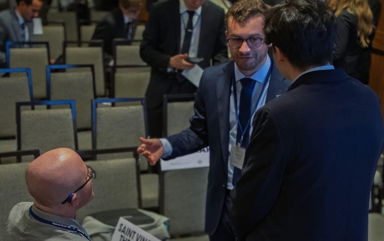 Ryleigh Newman, seen from behind with his Saint Vincent placard, talking with two standing delegates among rows of empty blue chairs during a committee break.